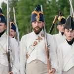 Soldiers marching in historical military uniforms reenact past events outdoors.