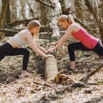 Side view of young female athletes in activewear stretching legs together using fallen tree during fitness workout in spring forest adhering to healthy lifestyle